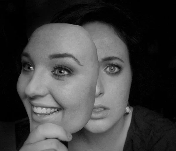 A woman holding a smiling mask of herself in front of her face, behind which she apparently feels very different. A woman holding a smiling mask of herself in front of her face, behind which she apparently feels very different.