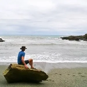 A man in a blue shirt and shorts with a head sitting on a big stone on an otherwise sandy beach, looking at the ocean A man in a blue shirt and shorts with a head sitting on a big stone on an otherwise sandy beach, looking at the ocean