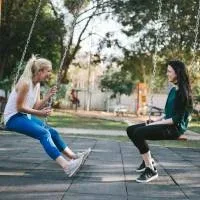 Two young women sitting on swings in a park, laughing together Two young women sitting on swings in a park, laughing together