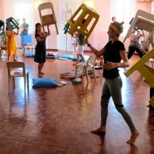 A room with several people practicing to balance a chair on one of its legs on their hand. A room with several people practicing to balance a chair on one of its legs on their hand.
